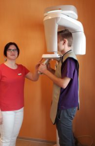 Das Bild zeigt eine Zahnarzthelferin, die bei einem Patienten ein Röntgenbild anfertigt. Der Patient ist im Röntgegerät positioniert.